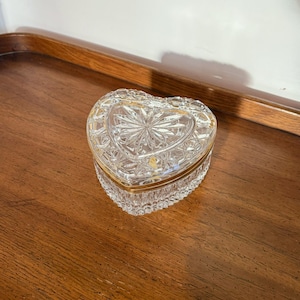 May include: A clear, heart-shaped trinket box with a gold-colored trim. The box is made of cut crystal, with a starburst design on the lid. The box is sitting on a wooden surface.