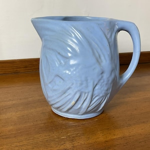 May include: A light blue ceramic pitcher with a textured, wavy design. The pitcher has a handle and a wide mouth.