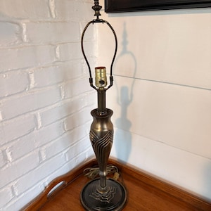May include: A bronze-toned table lamp with a decorative, fluted base and a circular foot. The lamp has a curved arm to hold a lampshade and a decorative finial at the top. The lamp is on a wooden tray.