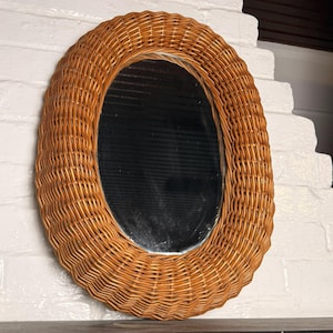 May include: An oval-shaped mirror featuring a thick, woven light brown wicker frame. The mirror has a smooth, reflective glass surface set within the textured, natural-colored rattan border, creating a distinct contrast between the glass and the weave.