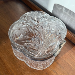 May include: A clear, four-lobed glass trinket box with a floral design etched on the lid. The box has a silver-colored metal hinge and sits on a wooden surface. The lid features etched flowers and leaves.