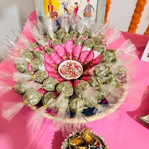 Puede incluir: Una presentación de pequeños obsequios envueltos, presentados en una cesta tejida y dispuestos sobre una superficie rosa. Los obsequios están rodeados de papel rosa y un letrero que dice "Treat Yourself to These Favors". Un pequeño cuenco de semillas mixtas está en el centro.