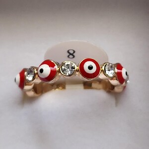 Puede incluir: Anillo dorado con diseños de ojo turco rojos y blancos alternados y gemas redondas y transparentes. El anillo es talla 8. Los símbolos del ojo turco son rojos con centro blanco y pupila negra. Las gemas son claras y facetadas.