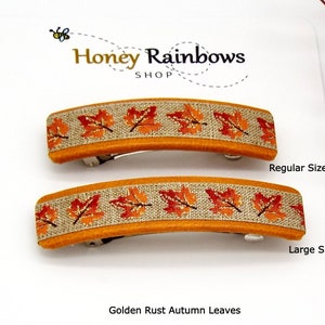 May include: Two hair clips with a golden rust orange border and a central strip featuring a repeating pattern of orange and red autumn leaves on a textured beige background. The clips are labeled as Regular Size and Large Size on the display card.