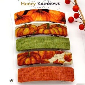 May include: A set of five rectangular barrettes. The barrettes feature various autumn-themed designs, including pumpkins and fall leaves. Colors include orange, green, and cream. The barrettes are made of fabric and have a metal clasp.