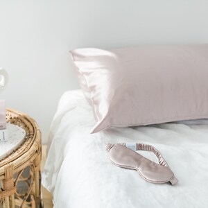 May include: A blush pink satin sleep mask and pillowcase on a white bed. The pillowcase is on a white bed with a white linen sheet. A small table with a white ceramic sculpture and a book titled "Alexa Chung" is next to the bed.