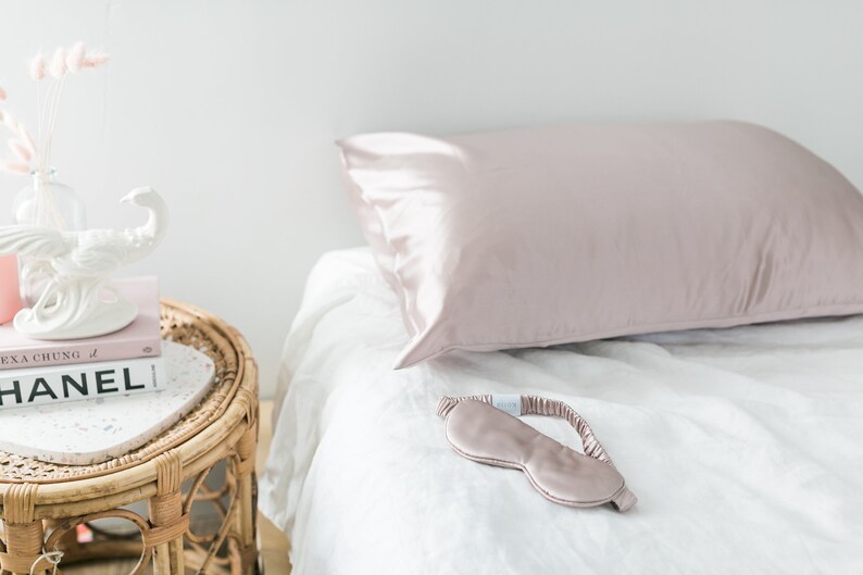 May include: A blush pink satin sleep mask and pillowcase on a white bed. The pillowcase is on a white bed with a white linen sheet. A small table with a white ceramic sculpture and a book titled "Alexa Chung" is next to the bed.