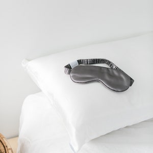 May include: A gray silk sleep mask with the word "QUION" printed on the strap, resting on a white pillow.