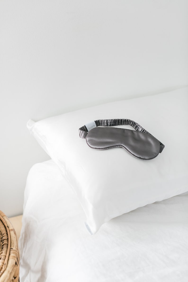 May include: A gray silk sleep mask with the word "QUION" printed on the strap, resting on a white pillow.