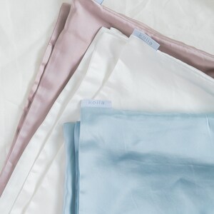 May include: Three different colored silk pillowcases, all with the same brand label, "koila", in a light blue font. The pillowcases are stacked on top of each other, with the pink pillowcase on top, the white pillowcase in the middle, and the light blue pillowcase on the bottom.