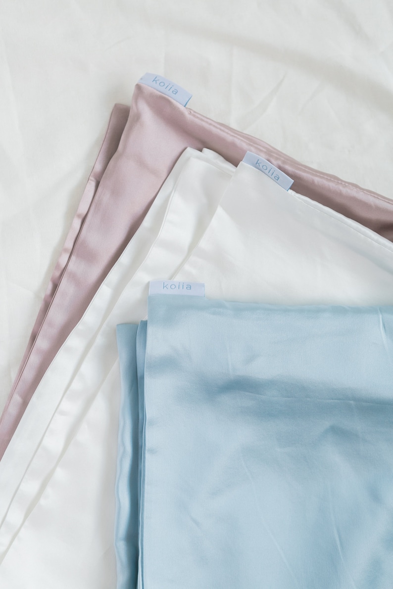 May include: Three different colored silk pillowcases, all with the same brand label, "koila", in a light blue font. The pillowcases are stacked on top of each other, with the pink pillowcase on top, the white pillowcase in the middle, and the light blue pillowcase on the bottom.