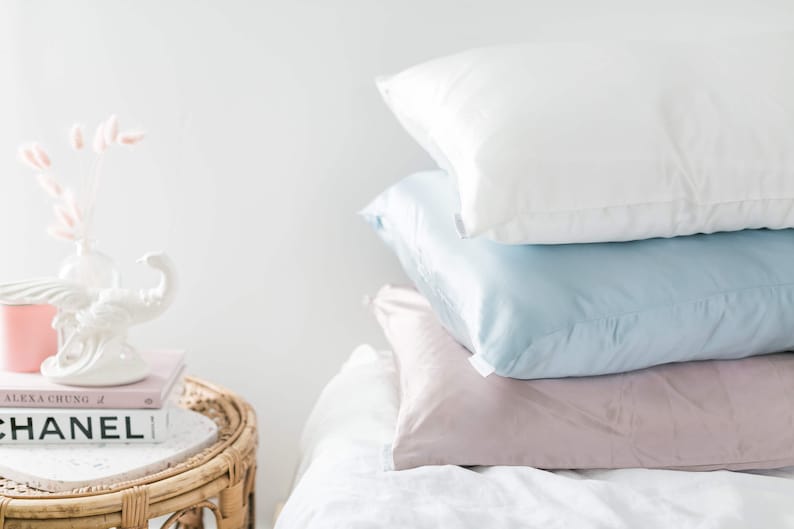 May include: Three pillows stacked on a white bed. The top pillow is white, the middle pillow is light blue, and the bottom pillow is light pink.
