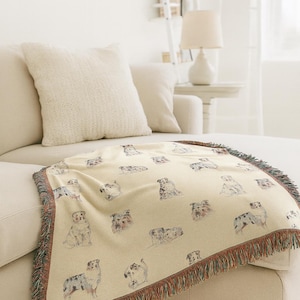 Peut inclure: Une couverture crème avec un motif répété de chiens gris et marron. La couverture a une bordure à franges marron, verte et rouge. Elle est drapée sur un canapé beige. Un élément de décoration intérieure.