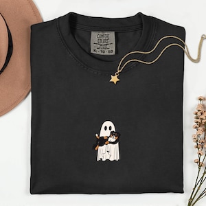 May include: Black crew neck t-shirt with a ghost and dog graphic. The ghost is white with black eyes and is holding a black, brown, and white dog. A gold star pendant necklace is draped across the shirt.