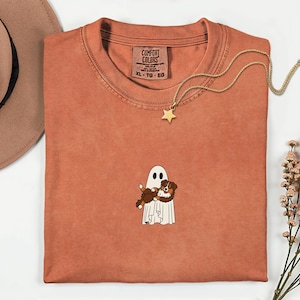 May include: Rust-coloured Comfort Colors t-shirt with a ghost and dog graphic. The ghost is holding the dog. A gold star pendant necklace is draped across the shirt. A brown hat is in the upper left corner.
