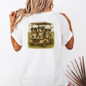 May include: White t-shirt featuring a watercolor illustration of three dogs in a golf cart. The dogs are wearing hats and appear to be enjoying a day on the golf course. The design is printed on the back of the shirt.