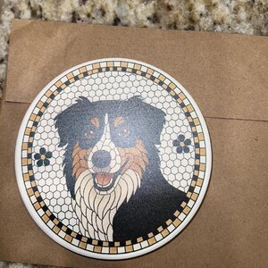 May include: A round ceramic coaster featuring a smiling dog with black, brown, and white fur. The dog is set against a mosaic-style background with a floral border. The coaster is on a brown paper surface.
