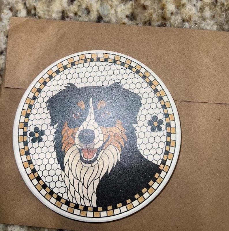 May include: A round ceramic coaster featuring a smiling dog with black, brown, and white fur. The dog is set against a mosaic-style background with a floral border. The coaster is on a brown paper surface.