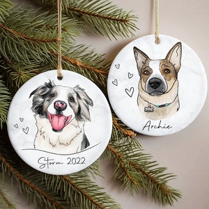 May include: Two white ceramic Christmas ornaments with dog portraits. The ornament on the left features a black and white dog with the name "Storm" and the year "2022" written below. The ornament on the right features a brown and white dog with the name "Archie" written below.
