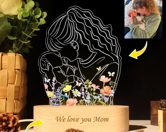 Puede incluir: Una silueta blanca de una madre sosteniendo un bebé, rodeada de flores coloridas. La base de la lámpara está hecha de madera y tiene el texto "We love you Mom" grabado. La lámpara está iluminada con una luz blanca cálida.