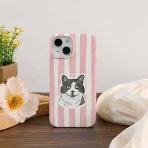 May include: A pink and white striped phone case featuring a cat illustration with the name "Zip". The cat is black and white with yellow eyes. The phone case has a camera and flash.