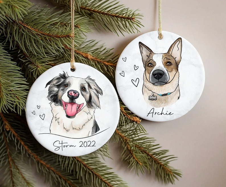 May include: Two white ceramic Christmas ornaments with dog portraits. The ornament on the left features a black and white dog with the name "Storm" and the year "2022" written below. The ornament on the right features a brown and white dog with the name "Archie" written below.