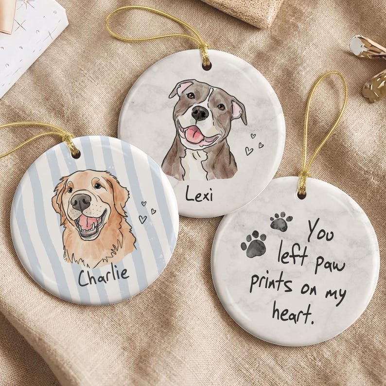May include: Three round ceramic ornaments with gold hanging strings. One features a golden retriever illustration labeled Charlie on blue stripes. Another shows a pit bull illustration labeled Lexi. The third has paw prints and the text You left paw prints on my heart.