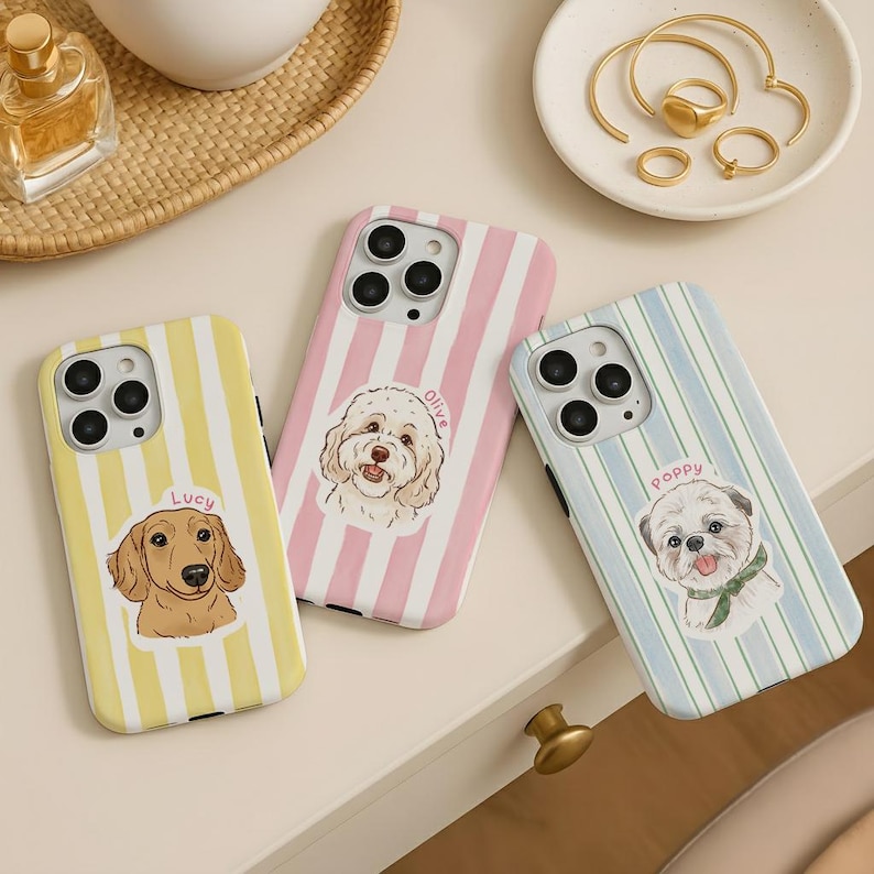May include: Three phone cases with dog illustrations. One case is yellow with vertical stripes and a dachshund illustration with the name "Lucy". Another is pink with vertical stripes and a dog illustration with the name "Olive". The third is blue with vertical stripes and a dog illustration with the name "Poppy".