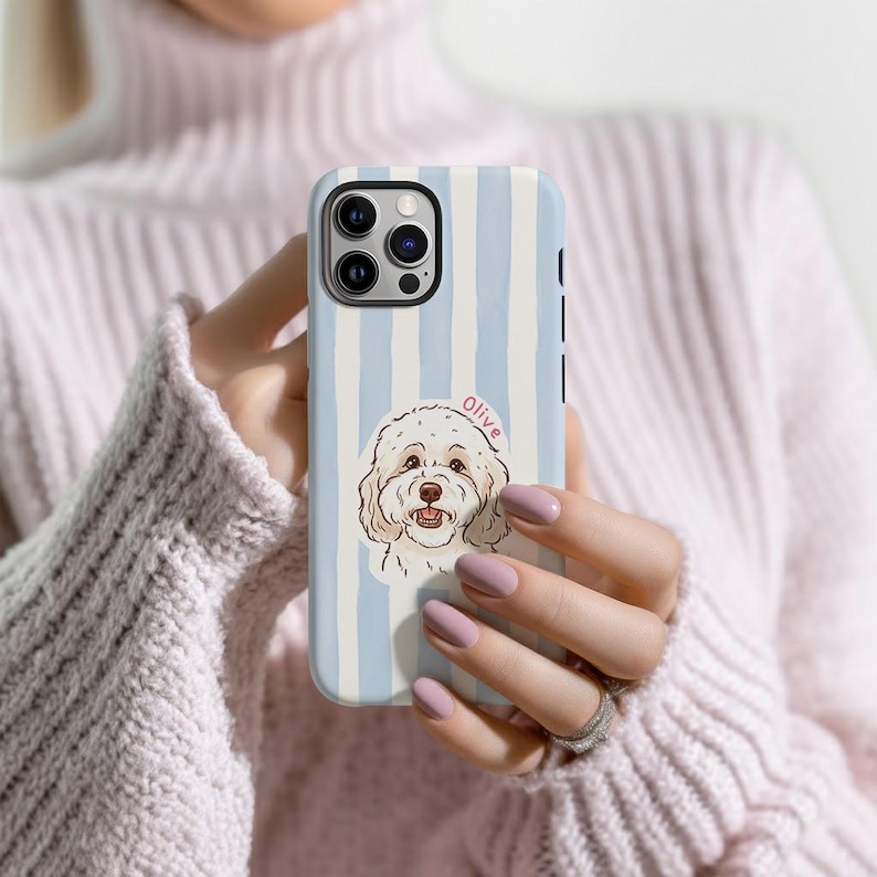 May include: A light blue and white striped phone case featuring a cartoon illustration of a dog with the name "Olive" in pink. The case has a triple-lens camera and black side buttons.