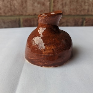 May include: A small, handmade ceramic bottle stopper. The stopper is a rich brown color with a glossy finish. It has a rounded base and a flared top, and is sitting on a white surface.