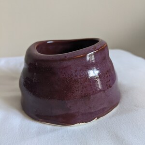 Pode incluir: Uma pequena tigela de cerâmica feita à mão com um esmalte roxo escuro. A tigela tem uma forma orgânica e assimétrica com uma borda ligeiramente alargada. O exterior tem uma aparência texturizada e o interior é liso. A base não é vidrada, revelando a cor natural da argila.