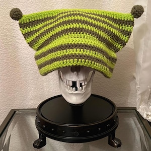 May include: A handmade crochet hat in a unique design. The hat features alternating stripes of lime green and dark brown. Two small brown pom-poms adorn the top. The hat is displayed on a white skull.