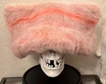 Könnte beinhalten: Eine weiße Totenkopfskulptur mit sichtbaren Zähnen wird von einem flauschigen, rosa und orange gestreiften Hut gekrönt. Der Schädel ruht auf einem schwarzen, verzierten Sockel mit rundem Oberteil und Klauenfüßen. Neutraler Hintergrund.