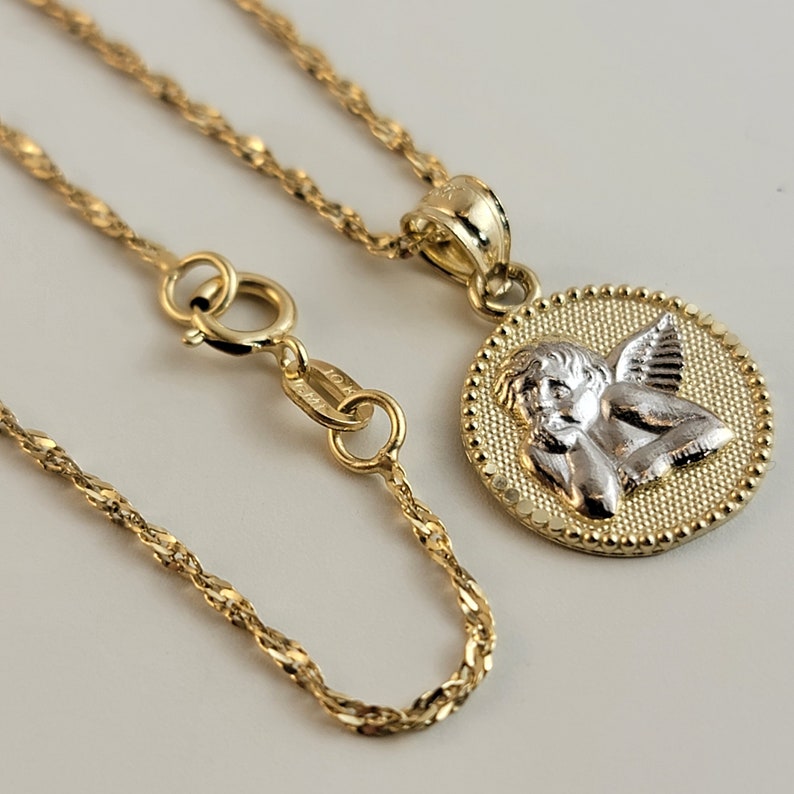 May include: A gold chain necklace with a round pendant featuring a detailed angel design. The angel is made of white gold and has wings and a halo. The pendant is surrounded by a beaded border.