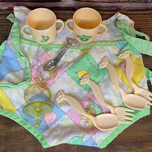May include: A vintage baby doll feeding set featuring two yellow cups, a silver spoon, and several cream-colored spoons and forks. The set includes a patchwork bib with a green trim and ties, and a pacifier.
