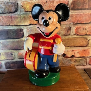Könnte beinhalten: Eine Vintage-Figur von Mickey Mouse in einer Marschkapellenuniform. Die Figur ist rot, blau und gelb und hält eine Trommel und einen Stab. Die Figur steht auf einem grünen Sockel.