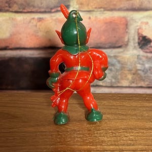 May include: A small, plastic Christmas elf ornament. The elf is red with green accents on the hat, shoes, and gloves. A gold string is attached for hanging. The elf is posed with its back to the viewer.