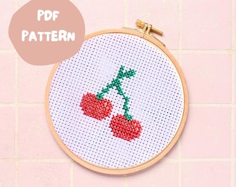 May include: A cross-stitch pattern of two red cherries with green stems and leaves on a white background. The pattern is in a wooden embroidery hoop.