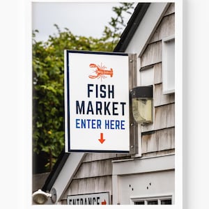 May include: A rectangular sign for a fish market, featuring a red lobster illustration at the top. The sign reads "FISH MARKET" in navy blue text, with "ENTER HERE" below, and a downward-pointing red arrow. The sign is mounted on a wooden building.