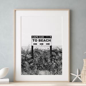 Peut inclure: Photographie en noir et blanc d'une pancarte indiquant "Cape Cod To Beach" avec des flèches pointant dans les deux sens. Le panneau est devant un bosquet de buissons.