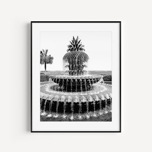 May include: A black and white photograph of a pineapple fountain with water flowing from the top. The fountain is surrounded by a circular pool of water. There is a palm tree in the background.