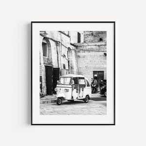 Puede incluir: Una fotografía en blanco y negro de un auto-rickshaw blanco estacionado en un callejón estrecho entre dos edificios de piedra. El auto-rickshaw está de frente a la cámara y tiene un conductor en el asiento. Hay otros vehículos estacionados en el fondo.