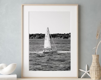 May include: Black and white photograph of a sailboat with a white sail on a body of water. The sailboat has the text "USA 187 TO" on the sail.