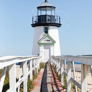 Vertical photograph of Brant Point Lighthouse in Nantucket, viewed straight down the weathered boardwalk with a bright blue sky and soft coastal light.