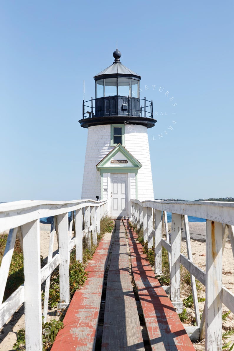 Vertical photograph of Brant Point Lighthouse in Nantucket, viewed straight down the weathered boardwalk with a bright blue sky and soft coastal light.