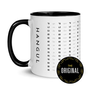 Puede incluir: Una taza de cerámica blanca con asa y borde negros. La taza presenta un gráfico en blanco y negro del alfabeto coreano, Hangul, con el texto "HANGUL" impreso verticalmente en el lateral. La taza también tiene una pegatina negra y amarilla con el texto "THE ORIGINAL" impreso en ella.