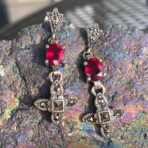 May include: A pair of silver-toned drop earrings with a star-shaped top, a red oval gemstone, and a cross-shaped bottom. The earrings are set with small, sparkling accents. The earrings are displayed on a textured, multi-colored surface.