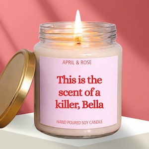 Op de afbeelding: Een brandende sojakaars in een glazen pot met een gouden deksel. Het etiket luidt "APRIL & ROSE" en "This is the scent of a killer, Bella". De kaars staat op een wit oppervlak tegen een roze achtergrond.