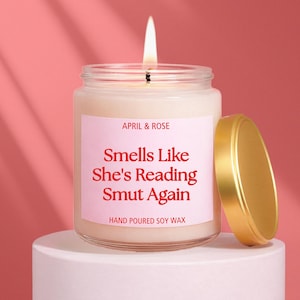 May include: A burning candle in a clear glass jar with a gold lid. The candle has a pink label with the text "APRIL & ROSE" and "Smells Like She's Reading Smut Again." The label also reads "HAND POURED SOY WAX."