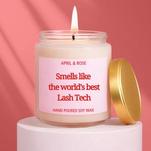 Könnte beinhalten: Eine brennende Kerze in einem durchsichtigen Glas mit goldenem Deckel. Auf dem Etikett steht "APRIL & ROSE" und "Smells like the world's best Lash Tech". Die Kerze steht auf einem weißen Sockel vor einem rosa Hintergrund. Der Text "HAND POURED SOY WAX" steht ebenfalls auf dem Etikett.